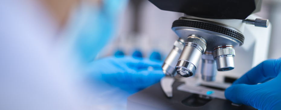 Immagine ravvicinata di un microscopio con una mano che regge un vetrino. L'operatore indossa guanti blu e si trova in un laboratorio, evidenziando un ambiente scientifico e di ricerca.