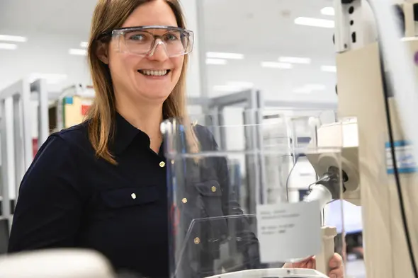 Donna sorridente in camice da laboratorio con occhiali protettivi, posizionata davanti a un laboratorio attrezzato. Sullo sfondo si intravedono strumenti e attrezzature da laboratorio.