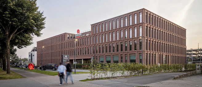 Immagine di un moderno edificio di mattoni con diverse finestre, situato lungo una strada. Alcune persone stanno camminando verso l'edificio, mentre auto sono parcheggiate nelle vicinanze. Sul lato dell'edificio è presente un'insegna luminosa.