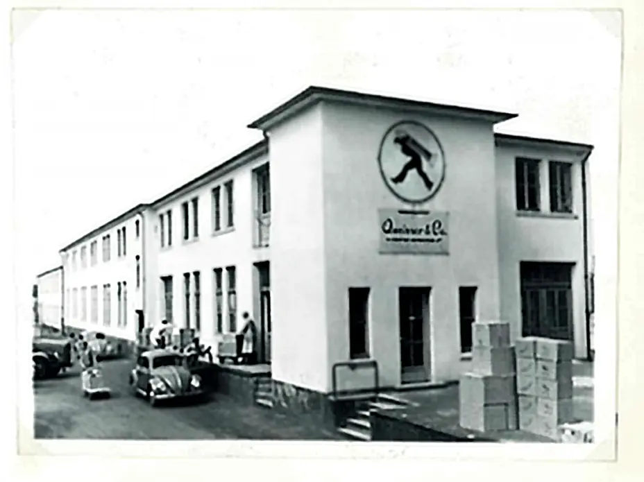 Immagine in bianco e nero di un edificio aziendale. La facciata presenta una grande insegna circolare con un simbolo al centro. Davanti si vedono alcune persone e auto d'epoca. Cassoni impilati sono visibili a lato dell'ingresso.