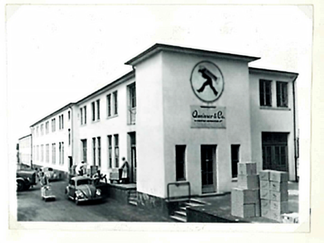 Immagine in bianco e nero di un edificio aziendale. La facciata presenta una grande insegna circolare con un simbolo al centro. Davanti si vedono alcune persone e auto d'epoca. Cassoni impilati sono visibili a lato dell'ingresso.