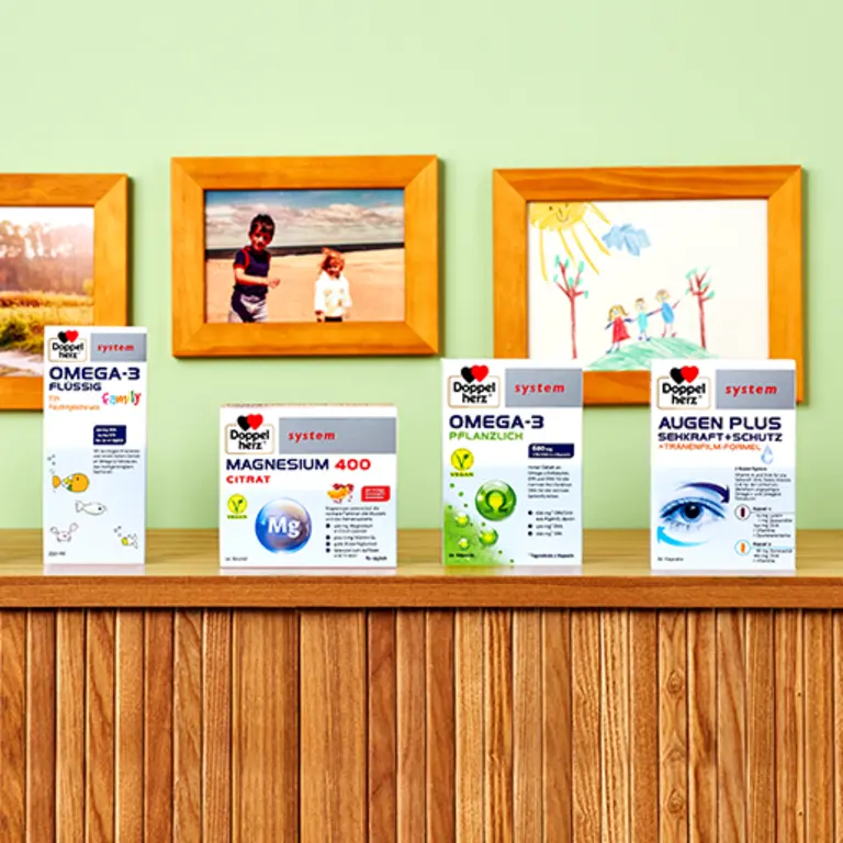Immagine di una base in legno con diversi prodotti salutistici Doppelt Herz sistemati in fila. I prodotti includono integratori di Omega-3, Magnesio 400 Citrato e Augen Plus, accanto a fotografie e disegni colorati su un muro verde chiaro.