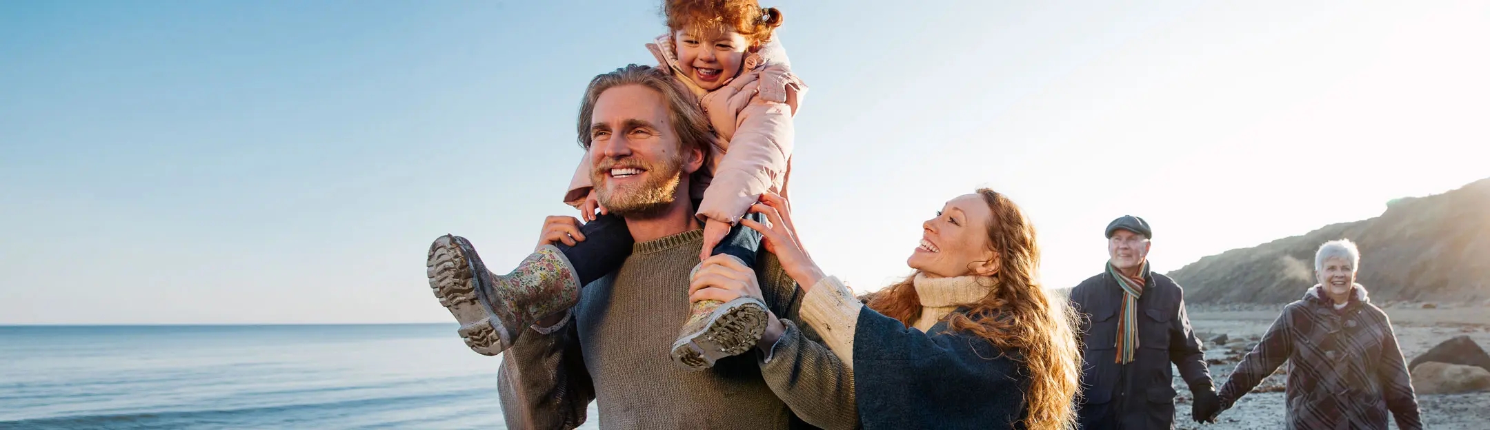 Una famiglia felice trascorre del tempo sulla spiaggia. Un uomo porta una bambina sulla spalle, mentre una donna lo accompagna sorridendo. Sullo sfondo, altre due persone camminano lungo la riva. Il mare è calmo e c'è una luce dorata al tramonto.