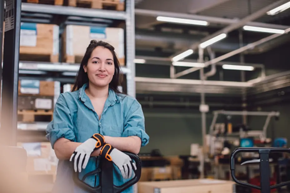 Una giovane donna sorridente indossa guanti da lavoro e si trova in un ambiente industriale, vicino a un carrello elevatore. Sullo sfondo si vedono scatole impilate e attrezzature di magazzino.