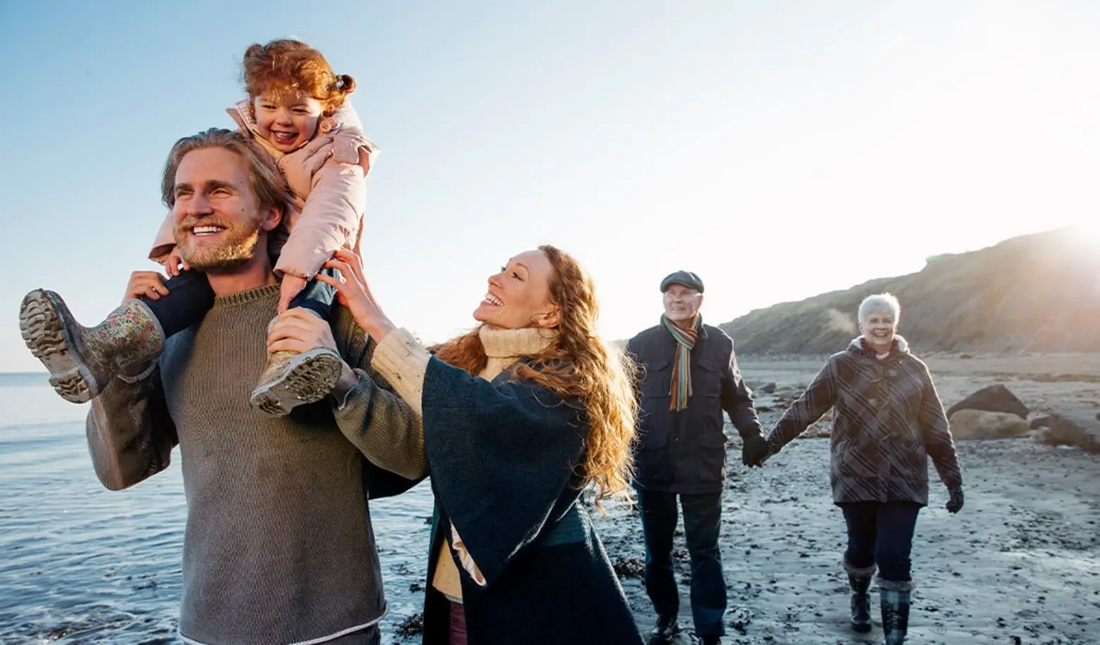 Una famiglia felice passeggia sulla spiaggia al tramonto. Un uomo sorregge una bambina sulle spalle, mentre una donna sorride accanto a lui. Un'altra coppia cammina mano nella mano sullo sfondo, godendo del momento insieme.