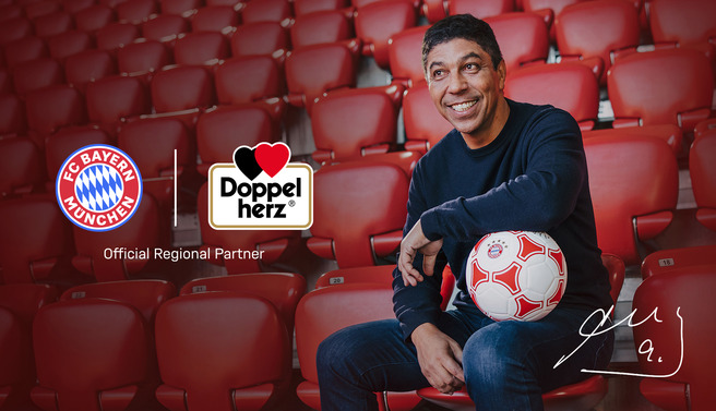 Un uomo sorridente è seduto sugli spalti di uno stadio, con una palla da calcio tra le mani. Dietro di lui ci sono i loghi del FC Bayern Monaco e di un'altra azienda, con la dicitura "Official Regional Partner".