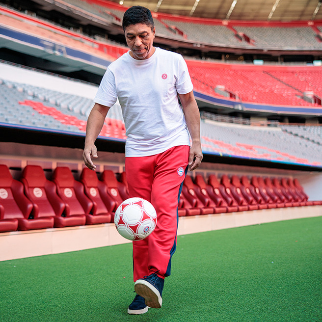 Un uomo in abbigliamento sportivo bianco e rosso sta dribblando un pallone da calcio. Si trova in uno stadio con seggiolini rossi sullo sfondo. L'atmosfera è di festa e sportività.