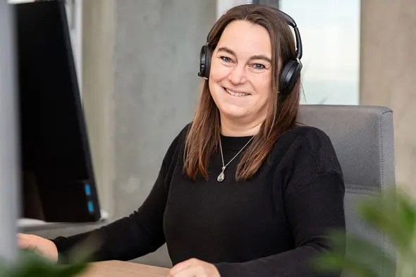 Donna sorridente con cuffie, seduta davanti a un computer, in un ambiente di lavoro moderno. Ha i capelli lunghi e castani e indossa un maglione nero. Si trova all'interno di un ufficio luminoso con pareti in vetro e piante in primo piano.