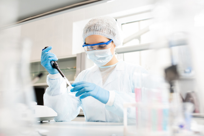Una persona in un laboratorio, vestita con un camice bianco, guanti, occhiali protettivi e mascherina, utilizza una pipetta per manipolare sostanze in un ambiente scientifico. Sullo sfondo si intravedono attrezzature da laboratorio.
