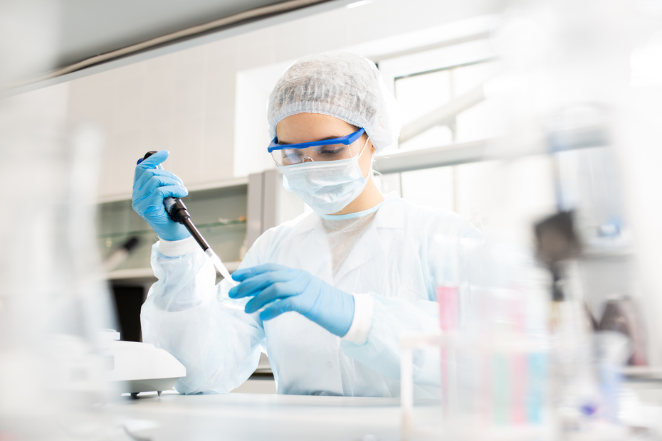 Una persona in un laboratorio, vestita con un camice bianco, guanti, occhiali protettivi e mascherina, utilizza una pipetta per manipolare sostanze in un ambiente scientifico. Sullo sfondo si intravedono attrezzature da laboratorio.