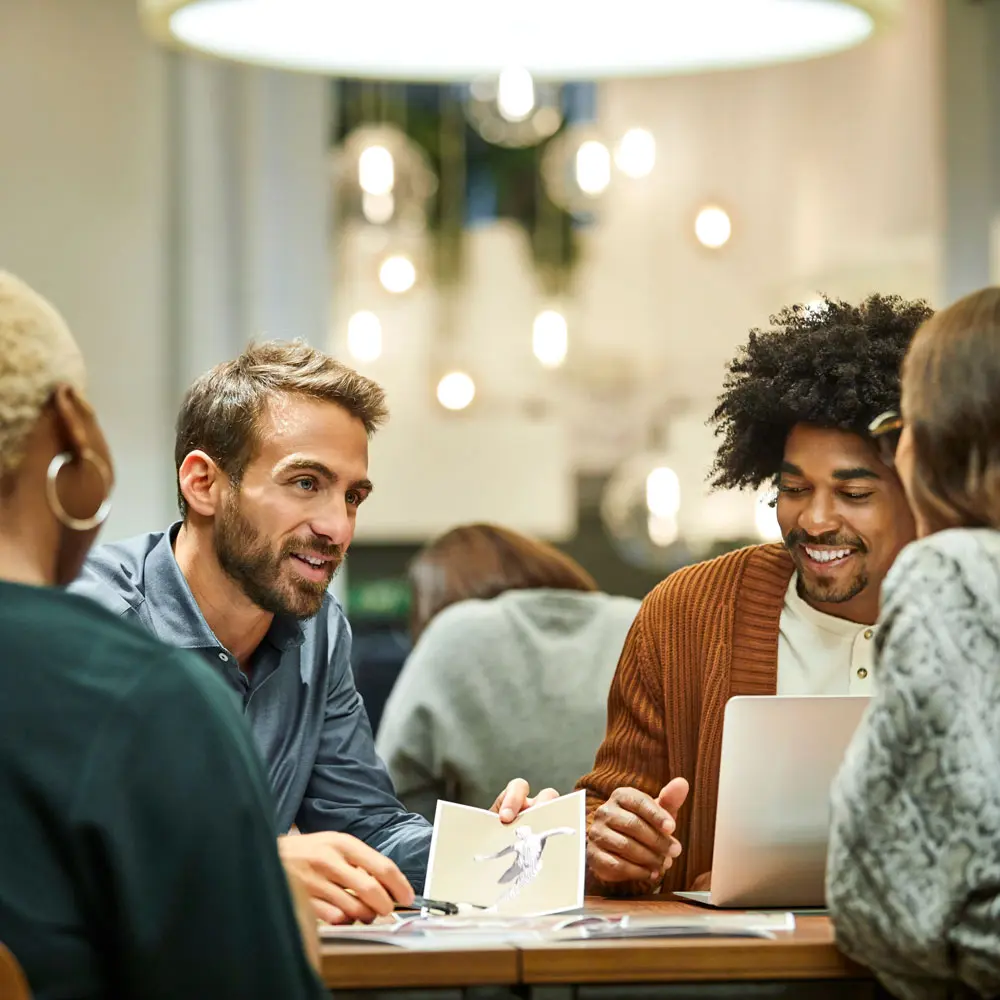 Un gruppo di persone è riunito attorno a un tavolo, discutendo. Un uomo con i capelli corti e una camicia blu interagisce con altri, mentre un altro uomo con i capelli ricci ascolta. L'atmosfera sembra informale e collaborativa.
