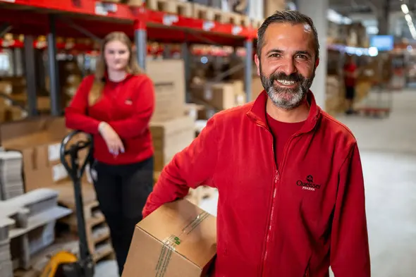 Due lavoratori in un magazzino, entrambi vestiti con maglioni rossi. Un uomo sorride mentre tiene una scatola, mentre una donna si trova sullo sfondo accanto a un carrello. Gli scaffali pieni di scatole sono visibili dietro di loro.