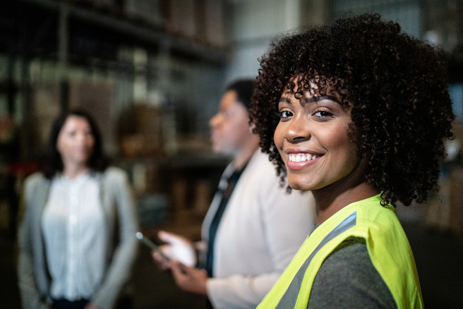 Un gruppo di tre persone in un ambiente di lavoro, probabilmente un magazzino. Una donna con capelli ricci e indossante un gilet di sicurezza sorride verso la fotocamera, mentre due donne discutono sullo sfondo. L'atmosfera appare professionale e collaborativa.