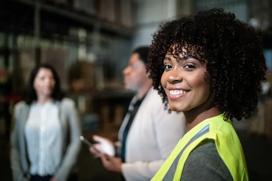 Un gruppo di tre persone in un ambiente di lavoro, probabilmente un magazzino. Una donna con capelli ricci e indossante un gilet di sicurezza sorride verso la fotocamera, mentre due donne discutono sullo sfondo. L'atmosfera appare professionale e collaborativa.