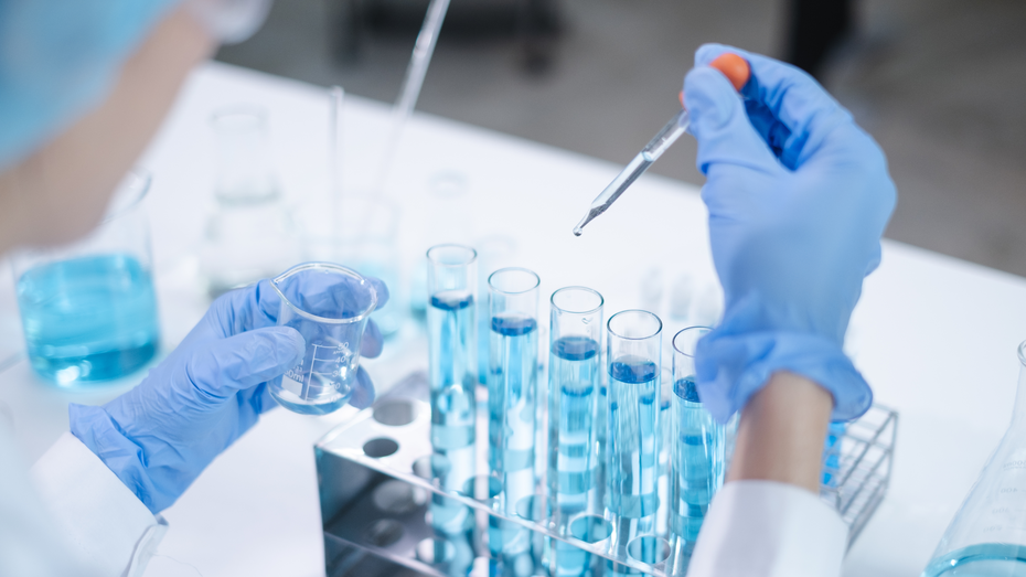 Un ricercatore in laboratorio indossa guanti blu e sta aggiungendo un liquido blu da una pipetta a provette simili. In un'altra mano tiene un contenitore con un altro campione. Sul tavolo ci sono diverse bottiglie e provette con liquido blu.