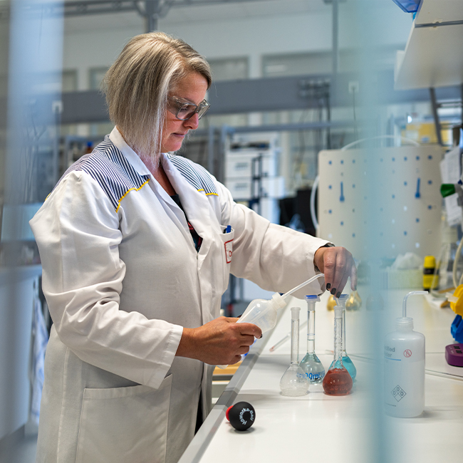 Una scienziata in un laboratorio indossa un camice bianco mentre utilizza strumenti di laboratorio. Di fronte a lei ci sono vari becher e cilindri con liquidi colorati, e il laboratorio è attrezzato con scaffali e strumenti scientifici.