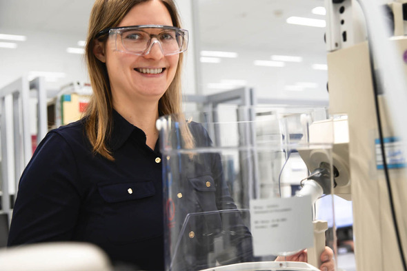 Donna sorridente in camice da laboratorio con occhiali protettivi, posizionata davanti a un laboratorio attrezzato. Sullo sfondo si intravedono strumenti e attrezzature da laboratorio.
